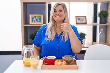 Sticker - Caucasian plus size woman eating breakfast at home doing happy thumbs up gesture with hand. approving expression looking at the camera showing success.