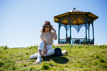 Happy young woman sitting on green grass. Dressed in white clothes and a hat. A walk in the fresh air on a sunny summer day