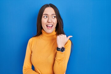 Canvas Print - Young brunette woman standing over blue background smiling with happy face looking and pointing to the side with thumb up.