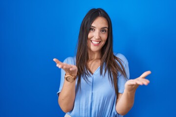 Poster - Young brunette woman standing over blue background smiling cheerful offering hands giving assistance and acceptance.