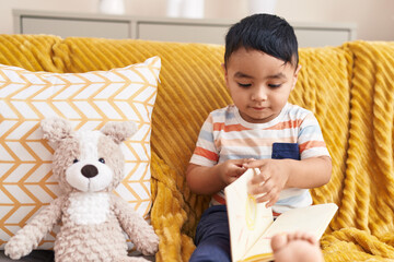 Wall Mural - Adorable hispanic toddler reading book sitting on sofa with teddy bear at home