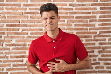 Canvas Print - Young hispanic man standing over bricks wall with hand on stomach because nausea, painful disease feeling unwell. ache concept.