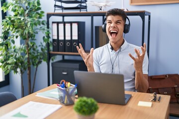 Sticker - Young hispanic man working at the office wearing headphones crazy and mad shouting and yelling with aggressive expression and arms raised. frustration concept.