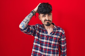 Poster - Young hispanic man with beard standing over red background confuse and wondering about question. uncertain with doubt, thinking with hand on head. pensive concept.