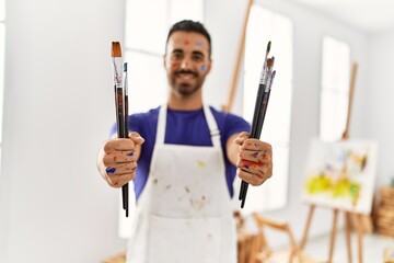 Sticker - Young hispanic man smiling confident holding paintbrushes at art studio