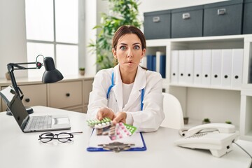 Sticker - Young brunette doctor woman holding pills making fish face with mouth and squinting eyes, crazy and comical.