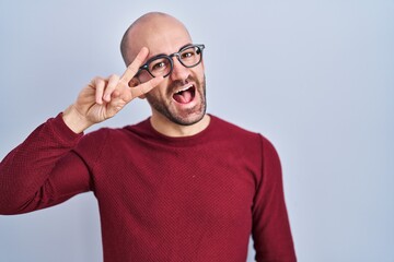 Poster - Young bald man with beard standing over white background wearing glasses doing peace symbol with fingers over face, smiling cheerful showing victory