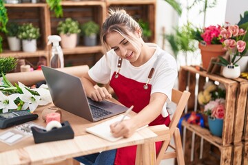 Wall Mural - Young blonde woman florist using laptop writing on notebook at flower shop