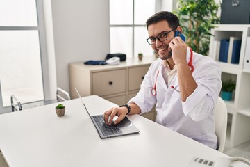 Wall Mural - Young hispanic man wearing doctor uniform using laptop talking on the smartphone at clinic