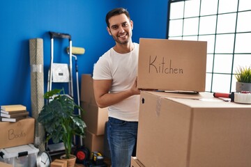 Wall Mural - Young hispanic man smiling confident holding kitchen cardboard box at new home