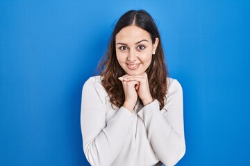 Sticker - Young hispanic woman standing over blue background laughing nervous and excited with hands on chin looking to the side