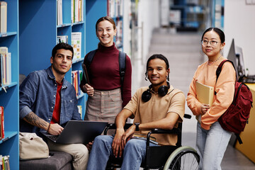 Wall Mural - Diverse group of students with young man in wheelchair looking at camera in college library, inclusivity concept