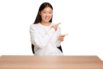 Wall Mural - Young asian woman sitting on a table isolated excited pointing with forefingers away.