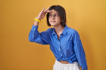 Poster - Young girl standing over yellow background very happy and smiling looking far away with hand over head. searching concept.