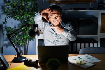 Canvas Print - Young chinese man working using computer laptop at night smiling making frame with hands and fingers with happy face. creativity and photography concept.