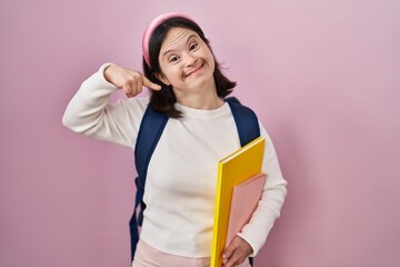 Poster - Woman with down syndrome wearing student backpack and holding books smiling cheerful showing and pointing with fingers teeth and mouth. dental health concept.