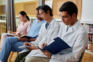 Group of medics in row taking notes during lecture or seminar, focus on young man holding clipboard in foreground