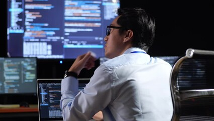 Close Up Back View Of Asian Male Programmer Thinking About Something While Writing Code By A Laptop Using Multiple Monitors Showing Database On Terminal Window Desktops In The Office
