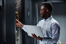Side View Portrait Of Young Black Man As Network Engineer Working With Servers In Data Center And Holding Laptop