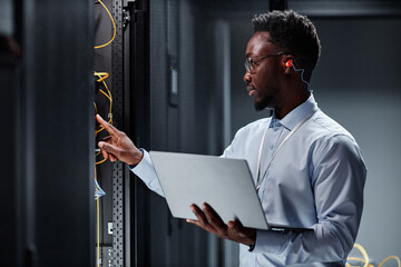 Side view portrait of young black man as network engineer working with servers in data center and holding laptop