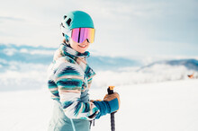 Woman In Skiing Clothes With Helmet And Ski Googles On Her Head With Ski Sticks. Winter Weather On The Slopes. Mountain And Enjoying View. Alpine Skier. Winter Sport. Ski Touring