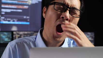 Close Up Of Asian Male Programmer Yawning And Sleeping While Writing Code By A Laptop Using Multiple Monitors Showing Database On Terminal Window Desktops In The Office
