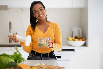 Wall Mural - Happy young african american woman making smoothie with blender at table with organic vegetables