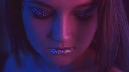 Neon light face. Party makeup. Nightclub beauty. Closeup portrait of purple blue color glow woman with closed eyes shimmering eyeshadow lips on dark.