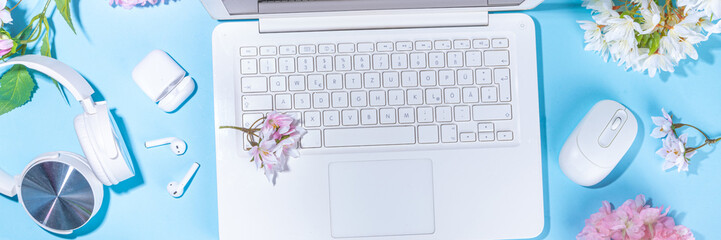 Spring office workplace, blogging flat lay background. White laptop, with headphones, tablet, spring flowers bouquet on light blue background, girl's hands typing on a laptop.