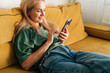 Caucasian middle aged woman browsing media in mobile phone while sitting on yellow sofa at home.