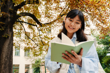 Wall Mural - Young asian girl reading book while sitting near the tree