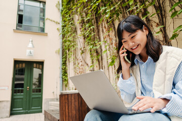 Wall Mural - Cute cheerful asian girl talking on mobile phone and working on laptop while sitting outdoors