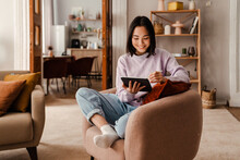 Young Smiling Asian Woman Drawing On Digital Tablet With Stylus While Sitting In Living Room