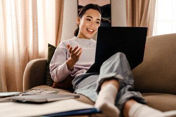 Wall Mural - Smiling asian woman making video call via laptop while sitting on couch