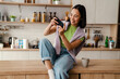 Cheerful asian woman playing online game on mobile phone while sitting in kitchen