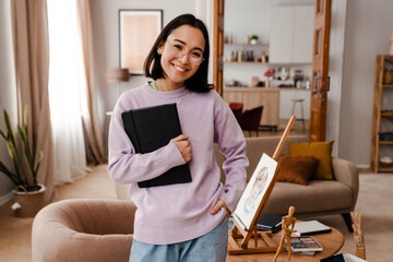 Wall Mural - Excited smiling asian woman artist standing in living room with sketchbook in hands