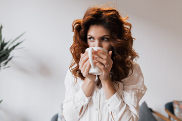 Wall Mural - Charming cute lovely woman with curly hair wearing white shirt is drinking coffee and looking aside while sitting in cafe. Woman dressed in white shirt is sitting on windowsill with cup of coffee. 