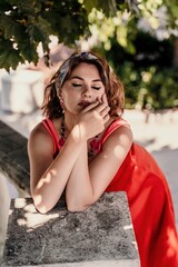 Wall Mural - A pretty woman in a red silk dress and a bandage on her head smiles against the background of the leaves of a tree. She is leaning on the coop and looking into the camera. Vertical photo.