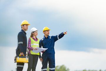 A team of 3 engineers, a man and a woman, are standing together pointing to an industrial construction project. and check the order wear a helmet Holding a blueprint and a toolbox and a blue uniform.