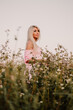 © Volodymyr - Vertical photo blonde woman posing in the big endless field of daisies in summer evening. Lady wear pink dressed, looking aside smiling, touching the plant green blossoming flower. Moody atmosphere