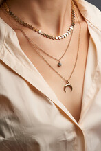 Cropped Close-up Portrait Of A Woman, Demonstrating Gold Necklaces With Pendants In A Beige Shirt. Set Of Three Different Chains. One Necklace Has A Crescent Moon Pendant.