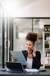 © Nuttapong punna - Business Africa woman Talking on the phone and using a laptop with a smile while sitting at modern office