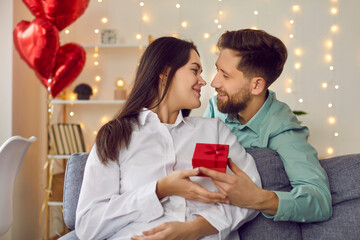 Wall Mural - Happy, handsome, young man gives gift to his attractive lover on Valentine's day, birthday, holiday. Young couple in love in romantic atmosphere are sitting on sofa in cozy living room at home.