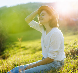 Wall Mural - Happy pretty young cheerful woman in white shirt enjoying sunny day while sitting on the green grass outdoor