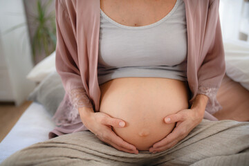 Wall Mural - Happy pregnant woman stroking her belly, sitting on bed.