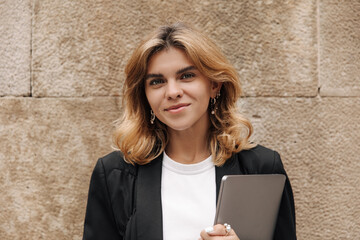 View of fair-haired young woman holding laptop and looking at camera. Smiling business lady in black jacket white t-shirt. Technology concept