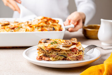 Poster - Serving, cooking homemade white lasagna with porcini and champignon mushrooms, onion and meat sausages. Pasta with parmesan cheese and bechamel sauce. Woman hands holding casserole.