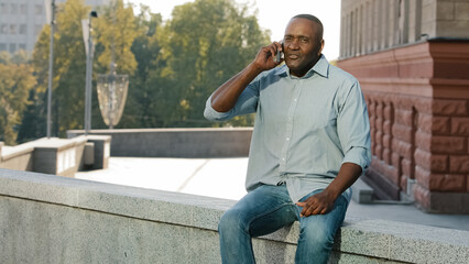 Adult mature African American man sitting outdoors holding cell phone angry middle-aged ethnic male businessman in city talking with smartphone has unpleasant mobile conversation unhappy with talk