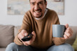 © LIGHTFIELD STUDIOS - angry man holding remote controller and cup of coffee in living room.
