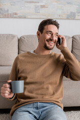 Wall Mural - smiling man in beige jumper talking on smartphone while sitting with cup of coffee at home.